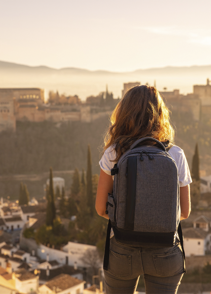 Persona en primer plano vista de espaldas, de pie en un mirador elevado, con una mochila gris de gran tamaño colocada sobre la espalda y vestimenta informal de tonos claros. Frente a la persona se extiende una ciudad histórica con edificaciones compactas, torres y cúpulas, parcialmente envuelta por una luz cálida de atardecer que crea un ambiente dorado y difuso. En el fondo se distinguen colinas suaves y una ligera bruma que atenúa los contornos del paisaje. En la parte inferior aparecen tejados y árboles altos y estrechos que aportan profundidad, mientras la iluminación suave resalta los volúmenes y genera una atmósfera tranquila y contemplativa.
