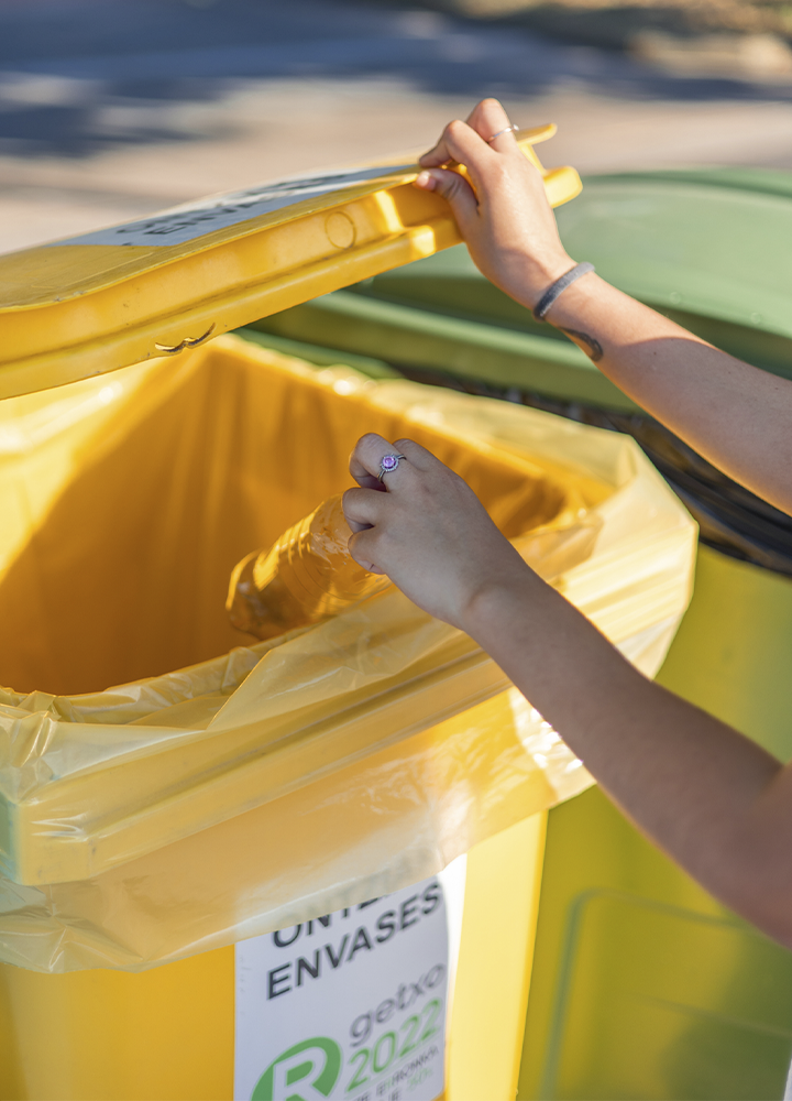 La imagen muestra una acción de reciclaje centrada en la correcta gestión de residuos de envases. En primer plano, unas manos depositan una botella de plástico vacía en un contenedor de color amarillo destinado a la recogida selectiva de envases, cuya tapa permanece abierta durante la acción. El contenedor está equipado con una bolsa interior amarilla y presenta una etiqueta visible en la que se puede leer parcialmente “ENVASES”. En segundo plano se observan otros contenedores de recogida selectiva, lo que refuerza el contexto de separación de residuos. La fotografía está tomada en un entorno exterior iluminado por luz natural y pone el foco en el gesto de reciclaje como práctica habitual de consumo responsable. En conjunto, la imagen transmite valores relacionados con la sostenibilidad, la economía circular, la reducción del impacto ambiental y el compromiso individual con la protección del medio ambiente mediante una adecuada gestión y reciclaje de los residuos.