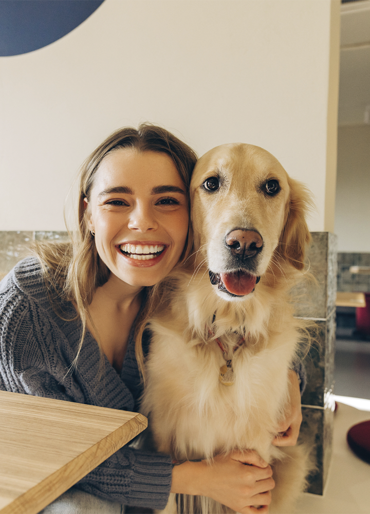 Persona abrazando a un perro de pelaje dorado en un interior luminoso, situados junto a una mesa de madera con un ordenador parcialmente visible en la esquina izquierda; al fondo se aprecian paredes claras con detalles geométricos circulares, una zona de paso y elementos decorativos en tonos neutros y cálidos, creando un ambiente moderno y acogedor.