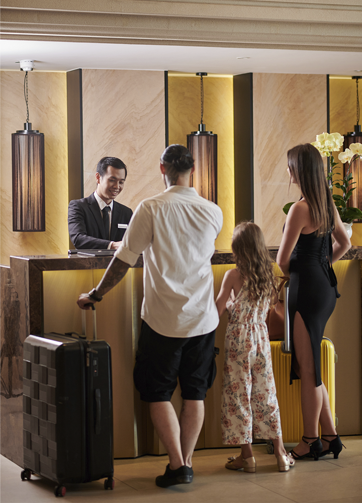 La imagen muestra el mostrador de recepción de un hotel, donde varias personas realizan el proceso de llegada o registro acompañadas de maletas de viaje. La escena se desarrolla en un vestíbulo elegante y cuidadosamente decorado, con acabados en tonos cálidos, iluminación ambiental y elementos ornamentales que reflejan un entorno acogedor y profesional. Detrás del mostrador, una persona del equipo de recepción atiende a los huéspedes mientras gestiona los trámites de entrada. Las maletas visibles refuerzan el contexto turístico y de alojamiento, asociando la imagen a la experiencia de acogida, atención al cliente y prestación de servicios hoteleros. En conjunto, la fotografía transmite valores de hospitalidad, calidad en el servicio y orientación a la satisfacción de las personas alojadas.
