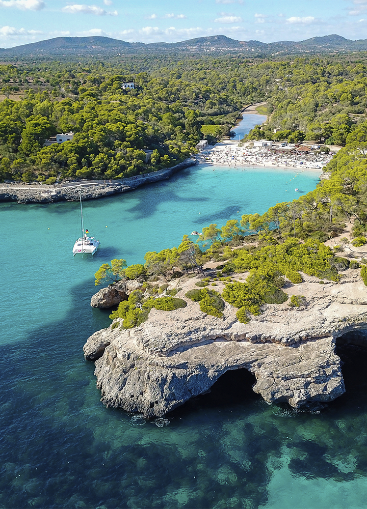 Vista aérea de una cala de agua turquesa rodeada de vegetación densa y colinas suaves al fondo; en primer plano, un acantilado rocoso con formas irregulares y pequeñas cuevas se adentra en el mar transparente. En el centro de la entrada a la cala hay un velero blanco fondeado, mientras al fondo se distingue una playa de arena clara con numerosas personas y zonas de sombra. El entorno natural combina tonos verdes de los pinos con azules intensos del agua, bajo un cielo despejado que ilumina la escena con luz natural y resalta la claridad del mar.