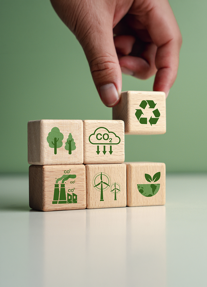 La imagen representa de forma simbólica los principales pilares de la sostenibilidad mediante una serie de bloques de madera ilustrados con iconos relacionados con el reciclaje, la protección de los recursos naturales, la reducción de emisiones de CO₂, las energías renovables y el cuidado del planeta. Una mano coloca la pieza superior, reflejando la importancia de la toma de decisiones y la acción responsable para construir un modelo de desarrollo más sostenible. En conjunto, la composición transmite el compromiso con la gestión ambiental, la economía circular y la integración de prácticas responsables que contribuyen a la protección del medio ambiente y al desarrollo sostenible.