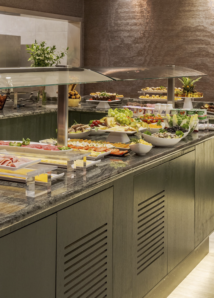 Buffet de autoservicio en un espacio interior de estilo moderno con iluminación cálida, donde una isla central de acabado oscuro y encimera de piedra sostiene bandejas y platos dispuestos de forma ordenada con alimentos variados como embutidos, frutas y ensaladas protegidos por una vitrina de cristal elevada con soportes metálicos; en el lateral y al fondo se observan más zonas de servicio con platos apilados, pequeños electrodomésticos, botellas y recipientes organizados, además de más bandejas con comida bajo otra cubierta; la pared trasera presenta un acabado texturizado en tonos marrones con focos de luz que crean reflejos suaves, y el suelo claro contrasta con el mobiliario oscuro, generando un ambiente limpio y cuidado orientado a la restauración.