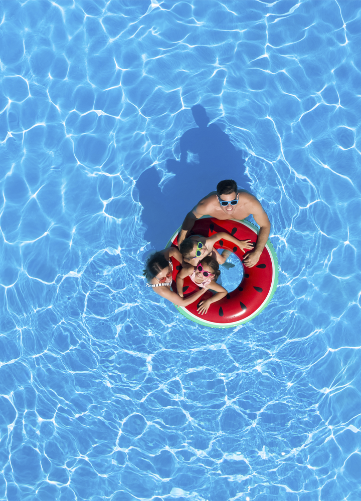 La imagen muestra una vista aérea de una piscina de agua cristalina en la que varias personas se encuentran sobre un flotador circular con diseño de sandía. La composición destaca por el amplio espacio ocupado por el agua azul, donde los reflejos de la luz generan patrones ondulados en toda la superficie. El flotador, de colores rojo, verde y negro, contrasta con el fondo acuático y se sitúa en la zona central de la imagen, convirtiéndose en el punto focal de la escena. En conjunto, la fotografía transmite una experiencia asociada al ocio, el disfrute de instalaciones recreativas y el tiempo libre en un entorno acuático, evocando actividades de descanso y convivencia durante la temporada estival.