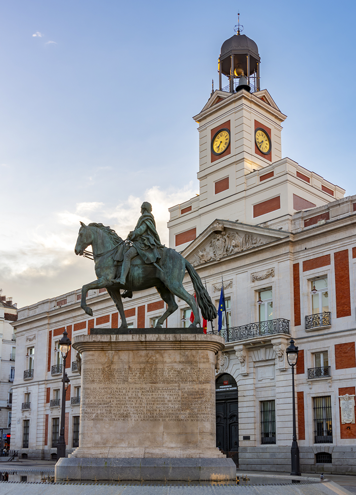 Estatua ecuestre de bronce situada sobre un pedestal de piedra en una plaza urbana, representando a una figura histórica montada a caballo en posición de avance. Detrás se alza un edificio institucional de fachada blanca y roja con balcones y un torreón central con reloj en cada cara, rematado por una estructura metálica en la parte superior. En la entrada principal se aprecian banderas, y a ambos lados hay farolas clásicas. La escena está iluminada con luz suave de atardecer, que resalta los volúmenes del edificio y la escultura sobre un cielo parcialmente nublado.