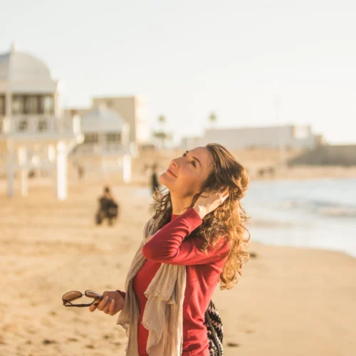 Persona de pie en una playa iluminada por la luz cálida del atardecer. Lleva una prenda roja y una bufanda clara, sostiene unas gafas de sol en una mano y se lleva la otra al cabello. El fondo aparece desenfocado, con arena, construcciones junto a la costa y algunas personas a lo lejos.