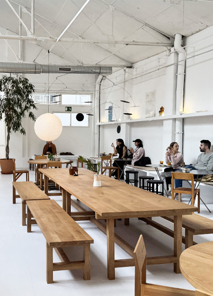 Espacio interior amplio y luminoso con estética minimalista, con techos altos blancos, vigas visibles y conductos expuestos. En primer plano hay una mesa larga de madera clara con bancos a ambos lados, sobre un suelo liso blanco. Al fondo, varias mesas pequeñas con personas sentadas, acompañadas de sillas de diseño sencillo y una iluminación colgante con formas esféricas y geométricas. En el lateral izquierdo aparece una planta alta en maceta. Las paredes son blancas con estanterías y objetos decorativos discretos, creando un ambiente de cafetería moderna o espacio de trabajo compartido.