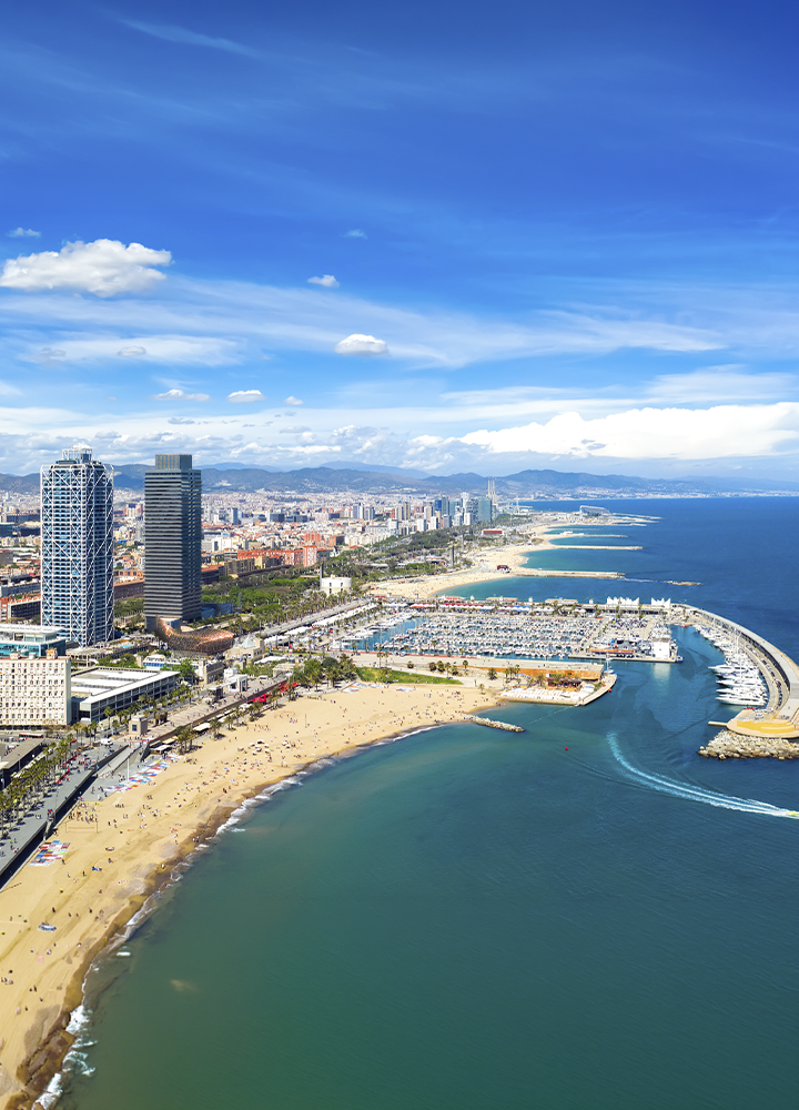 Vista aérea de una ciudad costera con una amplia playa de arena en primer plano curvada alrededor de una bahía, junto a un puerto deportivo lleno de embarcaciones protegido por diques; a la izquierda se elevan varios edificios altos y modernos cerca de la costa, mientras la ciudad se extiende hacia el fondo con construcciones más bajas, y el mar azul ocupa el lado derecho bajo un cielo amplio con nubes dispersas.