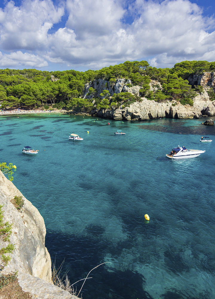 Bahía de aguas turquesa muy transparentes vista desde una zona rocosa en primer plano, con varias pequeñas embarcaciones flotando sobre el agua tranquila y algunas boyas amarillas dispersas; al fondo se alzan acantilados claros cubiertos de abundante vegetación verde y pinos, bajo un cielo parcialmente nublado con grandes nubes blancas que aportan contraste a la escena costera.