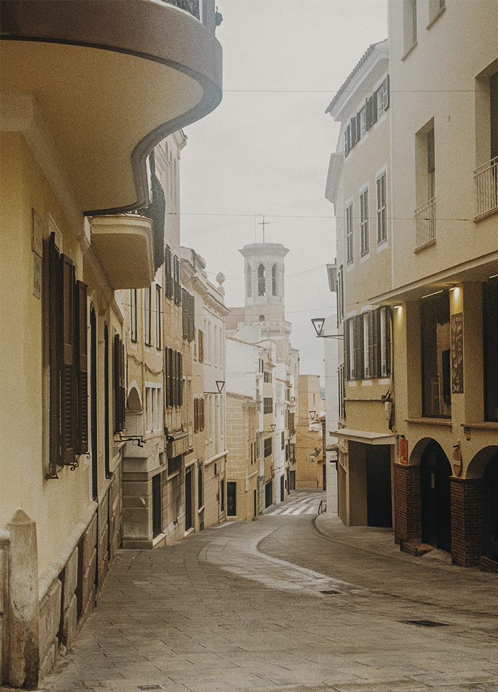 Calle estrecha y curvada de un entorno urbano tranquilo, flanqueada por edificios de varias plantas en tonos claros como beige y crema, con persianas oscuras y balcones pequeños; en el lado izquierdo destaca un balcón redondeado que sobresale sobre la vía, mientras que al fondo se eleva una torre clara con aberturas y una cruz en la parte superior. El pavimento de piedra y la ausencia de personas o vehículos refuerzan una atmósfera serena, bajo una luz suave y ligeramente difusa que aporta un aspecto cálido y atemporal.