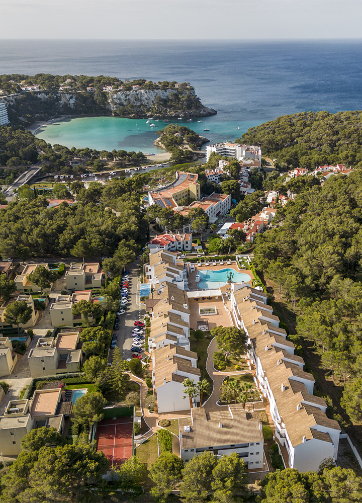 Vista aérea de una zona costera rodeada de abundante vegetación verde, con un complejo de edificios residenciales y hoteleros de tonos claros en primer plano organizados alrededor de caminos y una piscina central; más allá, una pequeña bahía de aguas turquesa queda protegida por formaciones rocosas y pinares, mientras el mar abierto se extiende hasta el horizonte bajo un cielo despejado, creando un contraste entre la arquitectura, el paisaje natural y el azul intenso del agua.