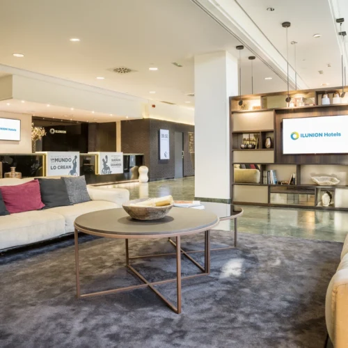 Vestíbulo moderno de un hotel ILUNION con zona de descanso compuesta por sofás beige, cojines decorativos y una mesa redonda sobre una alfombra gris. Al fondo se encuentra la recepción y una estantería con objetos decorativos y una pantalla que muestra el logotipo de ILUNION Hotels. El espacio está iluminado con luz cálida y combina tonos neutros, madera y detalles contemporáneos.