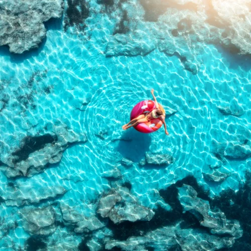 Vista cenital donde se encuentra con agua transparente de color turquesa; las olas suaves crean una franja de espuma blanca en la transición. Sobre el agua, una persona está recostada en un flotador circular de color rosa brillante, con las piernas extendidas hacia el centro del aro y el cuerpo en posición relajada. En la parte inferior se perciben rocas sumergidas bajo la superficie clara, y la luz del sol genera patrones ondulantes en el fondo marino, aportando sensación de calma y verano.