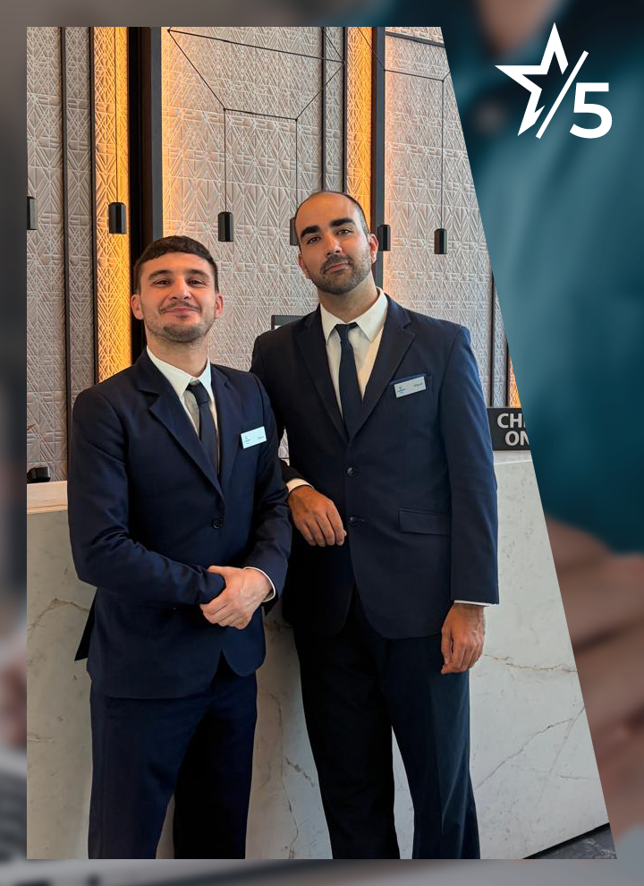 Dos personas con traje oscuro y corbata posan juntas en el interior de una recepción, situadas frente a un mostrador de piedra clara; una de ellas apoya el brazo sobre la encimera mientras la otra mantiene las manos entrelazadas delante. Detrás se observa una pared decorativa con paneles de textura geométrica y luces cálidas verticales, además de varios apliques negros colgantes. En la esquina superior derecha aparece un gráfico con una estrella y el número “5”.
