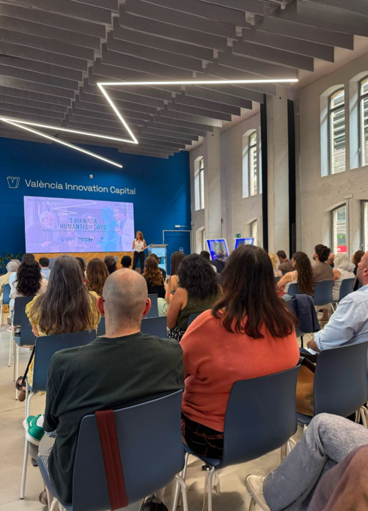 Sala amplia y luminosa con techo alto de vigas blancas y luces lineales modernas, donde varias filas de sillas ocupadas por asistentes miran hacia un escenario con pantalla; al fondo se lee “València Innovation Capital” sobre una pared azul mientras una persona presenta frente al público.