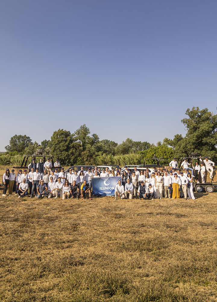 Grupo numeroso de personas posando en un campo seco al aire libre, organizadas en varias filas y vestidas mayoritariamente con ropa clara, con una pancarta azul de “ILUNION” en el centro; a ambos lados se observan vehículos tipo remolque con más personas subidas, y al fondo aparecen árboles verdes bajo un cielo despejado de gran amplitud.