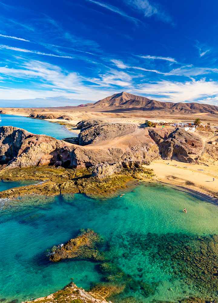 Costa volcánica con aguas transparentes de tonos verdes y azules, pequeñas calas entre formaciones rocosas y arena dorada; al fondo, una montaña árida bajo un cielo intenso con nubes alargadas.