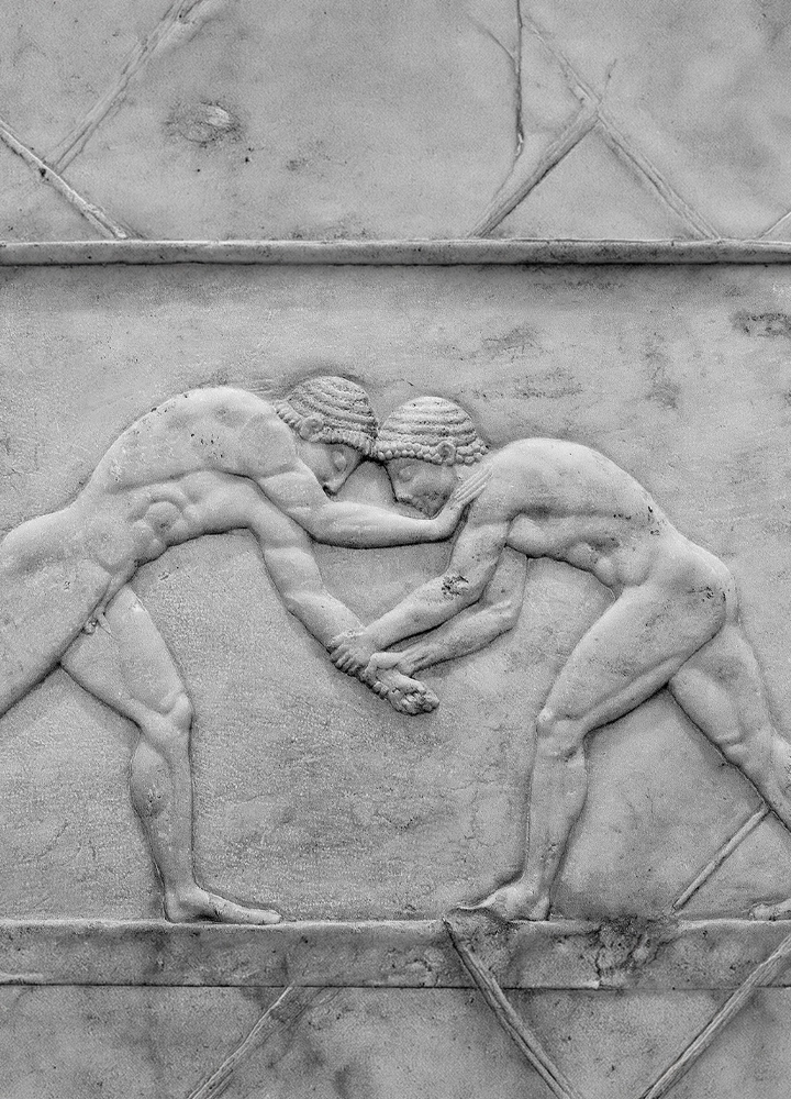 Relieve escultórico en piedra que representa a dos figuras humanas desnudas enfrentadas en una escena de lucha. Ambas aparecen inclinadas hacia delante, con las cabezas juntas y las manos entrelazadas mientras intentan sujetarse mutuamente. Los cuerpos muestran una musculatura marcada y están tallados en bajo relieve sobre una superficie de piedra clara con patrones geométricos grabados alrededor. La composición es simétrica y recuerda a una representación clásica de competición deportiva o lucha grecorromana.