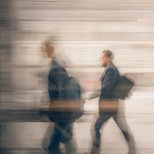 Fotografía con efecto de movimiento en la que dos personas caminan por un espacio urbano o de tránsito interior. Ambas llevan mochilas o bolsas al hombro, mientras el fondo aparece difuminado por una exposición prolongada que crea líneas horizontales y una sensación de velocidad. Predominan los tonos grises y beige, con las figuras destacando ligeramente sobre un entorno desenfocado.