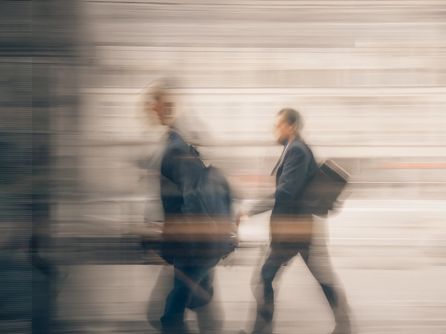 Fotografía con efecto de movimiento en la que dos personas caminan por un espacio urbano o de tránsito interior. Ambas llevan mochilas o bolsas al hombro, mientras el fondo aparece difuminado por una exposición prolongada que crea líneas horizontales y una sensación de velocidad. Predominan los tonos grises y beige, con las figuras destacando ligeramente sobre un entorno desenfocado.