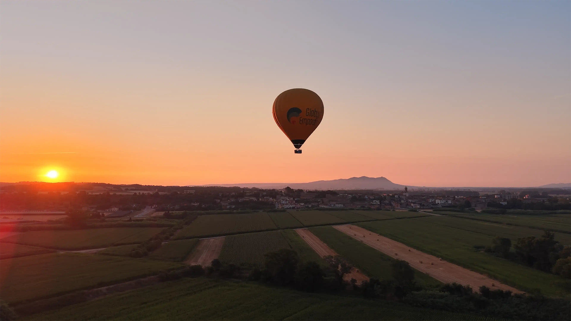 Globo aerostático volando a baja altura sobre un paisaje rural al atardecer, con campos verdes y parcelas agrícolas extendiéndose en primer plano y un pequeño núcleo urbano al fondo; el sol se sitúa cerca del horizonte a la izquierda, proyectando una luz cálida anaranjada que ilumina el cielo despejado y realza las montañas suaves en la distancia, mientras el globo queda centrado en la escena como punto focal.