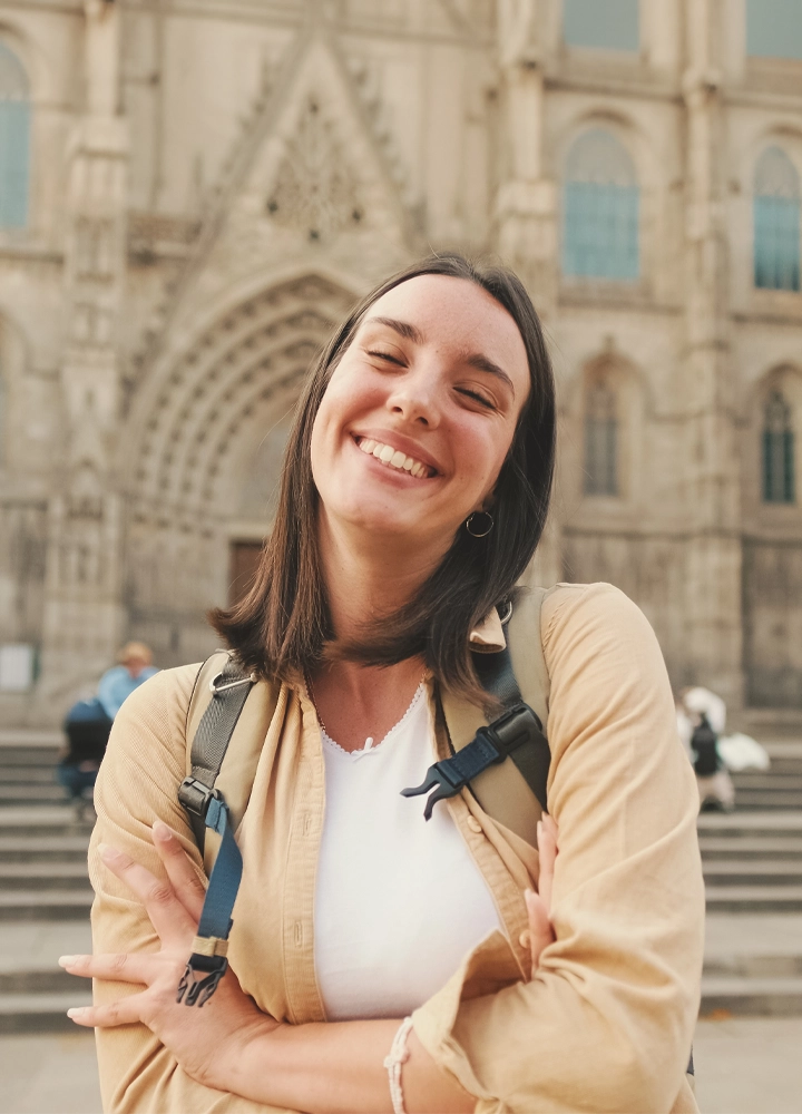Persona con mochila posa frente a la fachada de una catedral de estilo gótico. Aparece en primer plano con los brazos cruzados, mientras al fondo se distinguen las escalinatas y los detalles arquitectónicos del edificio histórico, ligeramente desenfocados. La iluminación cálida resalta los tonos beige de la ropa y la piedra del entorno.
