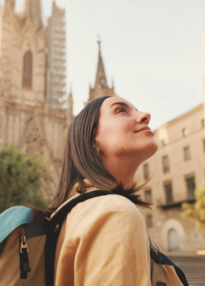 Persona con mochila observando hacia arriba en un entorno urbano histórico. Al fondo se alzan las torres de una catedral desenfocada y edificios de piedra iluminados por una luz cálida, creando una atmósfera de viaje y descubrimiento.