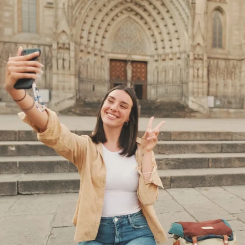 Persona sentada en una plaza frente a la fachada de una catedral gótica, tomando un selfi con un teléfono móvil. Con una mano sostiene el móvil y con la otra hace el gesto de la victoria. En primer plano se ve una mochila apoyada en el suelo, mientras al fondo destacan las escalinatas y el gran portal ornamentado del edificio histórico.