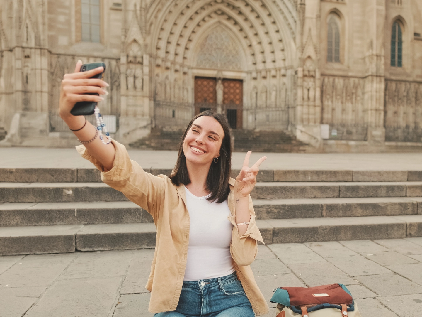 Persona sentada en una plaza frente a la fachada de una catedral gótica, tomando un selfi con un teléfono móvil. Con una mano sostiene el móvil y con la otra hace el gesto de la victoria. En primer plano se ve una mochila apoyada en el suelo, mientras al fondo destacan las escalinatas y el gran portal ornamentado del edificio histórico.