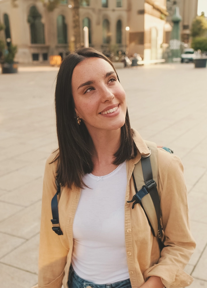 Persona con mochila en una plaza urbana de estilo histórico. En primer plano aparece mirando hacia arriba, mientras al fondo se distinguen edificios de piedra y una amplia zona peatonal iluminada por una luz cálida. La profundidad de campo desenfoca el entorno y centra la atención en la figura.