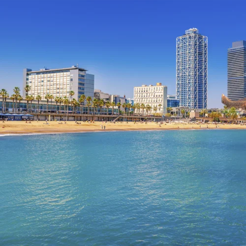 Vista de una playa urbana con arena clara en primer plano junto a agua tranquila de tono azul verdoso, donde varias personas se distribuyen a lo largo de la orilla; detrás se alinea un paseo con palmeras y, a continuación, varios edificios de diferentes alturas y estilos, incluyendo una torre alta de estructura moderna y un hotel de fachada clara, todo bajo un cielo completamente despejado de color azul intenso.
