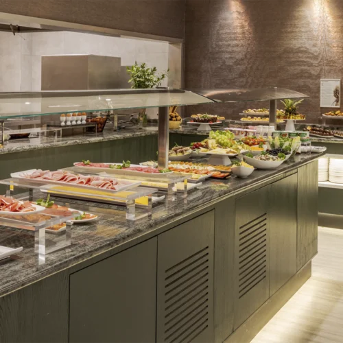 Buffet de autoservicio en un espacio interior de estilo moderno con iluminación cálida, donde una isla central de acabado oscuro y encimera de piedra sostiene bandejas y platos dispuestos de forma ordenada con alimentos variados como embutidos, frutas y ensaladas protegidos por una vitrina de cristal elevada con soportes metálicos; en el lateral y al fondo se observan más zonas de servicio con platos apilados, pequeños electrodomésticos, botellas y recipientes organizados, además de más bandejas con comida bajo otra cubierta; la pared trasera presenta un acabado texturizado en tonos marrones con focos de luz que crean reflejos suaves, y el suelo claro contrasta con el mobiliario oscuro, generando un ambiente limpio y cuidado orientado a la restauración.