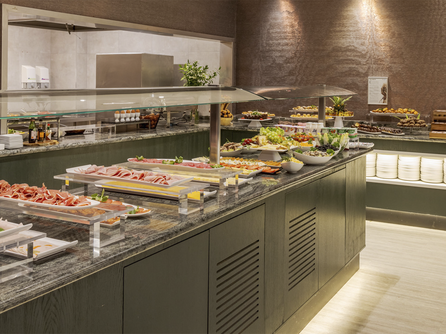 Buffet de autoservicio en un espacio interior de estilo moderno con iluminación cálida, donde una isla central de acabado oscuro y encimera de piedra sostiene bandejas y platos dispuestos de forma ordenada con alimentos variados como embutidos, frutas y ensaladas protegidos por una vitrina de cristal elevada con soportes metálicos; en el lateral y al fondo se observan más zonas de servicio con platos apilados, pequeños electrodomésticos, botellas y recipientes organizados, además de más bandejas con comida bajo otra cubierta; la pared trasera presenta un acabado texturizado en tonos marrones con focos de luz que crean reflejos suaves, y el suelo claro contrasta con el mobiliario oscuro, generando un ambiente limpio y cuidado orientado a la restauración.