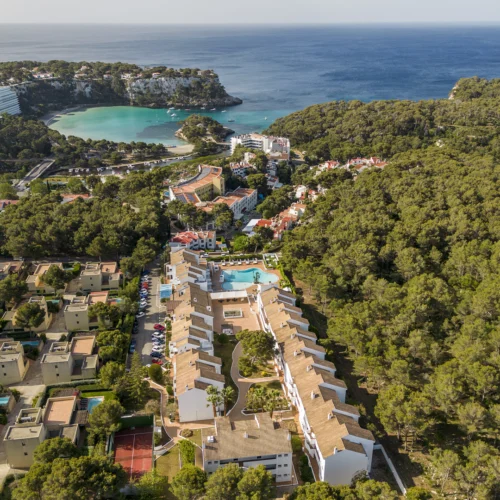 Vista aérea de una zona costera rodeada de abundante vegetación verde, con un complejo de edificios residenciales y hoteleros de tonos claros en primer plano organizados alrededor de caminos y una piscina central; más allá, una pequeña bahía de aguas turquesa queda protegida por formaciones rocosas y pinares, mientras el mar abierto se extiende hasta el horizonte bajo un cielo despejado, creando un contraste entre la arquitectura, el paisaje natural y el azul intenso del agua.
