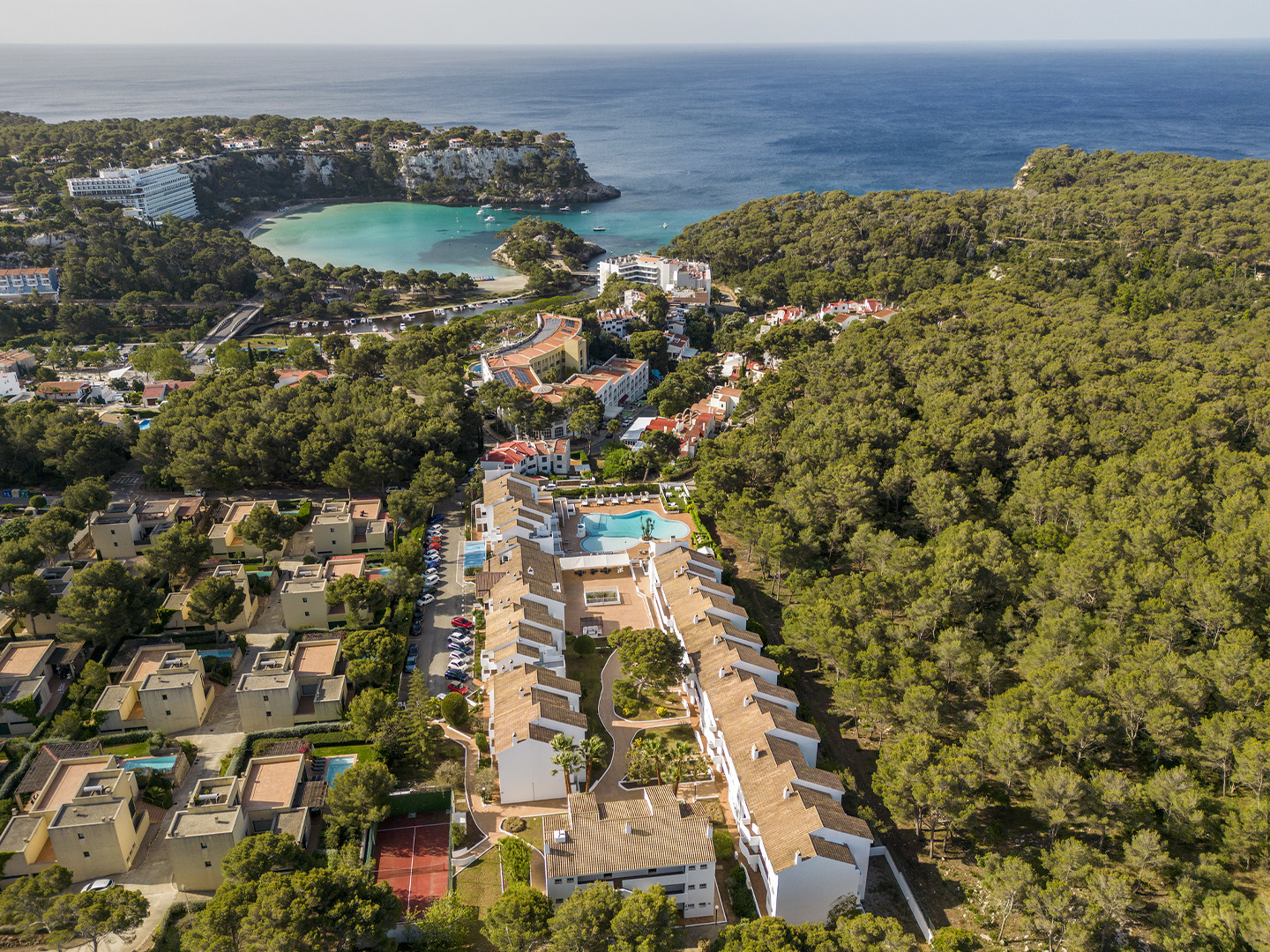 Vista aérea de una zona costera rodeada de abundante vegetación verde, con un complejo de edificios residenciales y hoteleros de tonos claros en primer plano organizados alrededor de caminos y una piscina central; más allá, una pequeña bahía de aguas turquesa queda protegida por formaciones rocosas y pinares, mientras el mar abierto se extiende hasta el horizonte bajo un cielo despejado, creando un contraste entre la arquitectura, el paisaje natural y el azul intenso del agua.