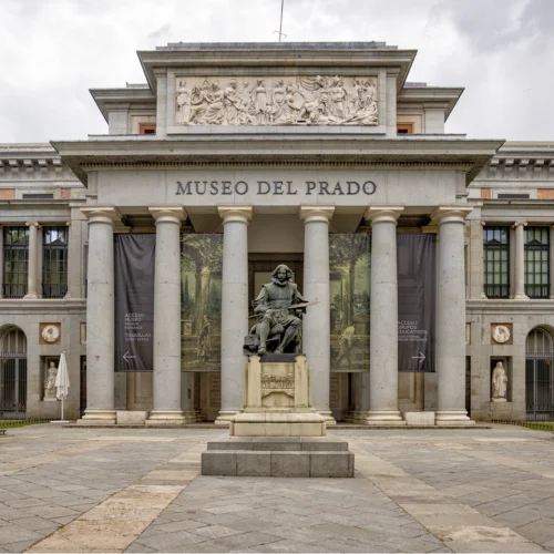 Fachada principal del Museo del Prado construida en piedra clara con un pórtico de columnas clásicas que sostienen un frontón decorado con relieves escultóricos de figuras humanas en movimiento, en cuyo centro se lee en letras grandes “MUSEO DEL PRADO”; frente a la entrada se alza una estatua de un personaje sentado sobre un pedestal, con un objeto alargado apoyado sobre las piernas, situada sobre una escalinata de piedra, mientras a ambos lados de la puerta cuelgan grandes lonas verticales con imágenes y texto relacionado con el acceso al museo, todo bajo una luz suave que resalta la textura de la arquitectura y el tono beige del edificio.