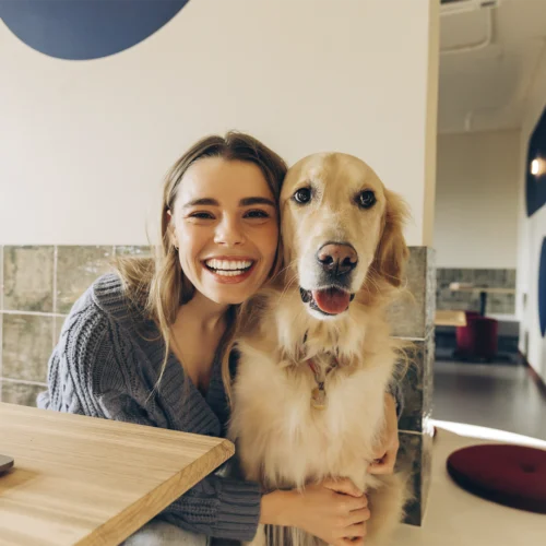 Persona abrazando a un perro de pelaje dorado en un interior luminoso, situados junto a una mesa de madera con un ordenador parcialmente visible en la esquina izquierda; al fondo se aprecian paredes claras con detalles geométricos circulares, una zona de paso y elementos decorativos en tonos neutros y cálidos, creando un ambiente moderno y acogedor.