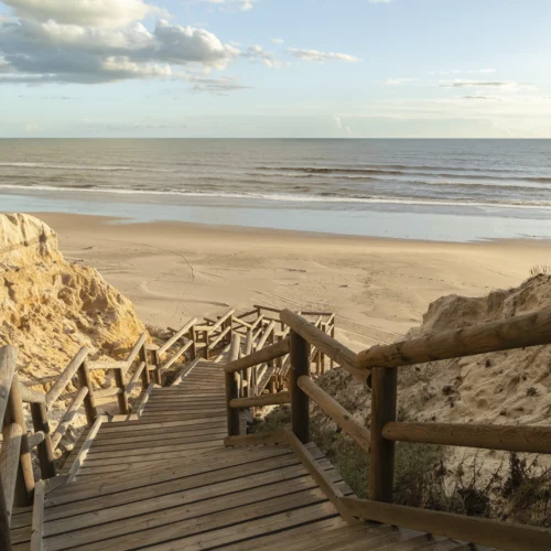 Acceso a una playa desde una pasarela de madera con escalones y barandillas en ambos lados, descendiendo entre formaciones de arena elevadas que crean pequeñas dunas a izquierda y derecha; la estructura guía la mirada hacia la orilla, donde se extiende una amplia superficie de arena lisa que llega hasta el mar, con olas suaves y paralelas acercándose al litoral; el horizonte divide el mar y un cielo parcialmente nublado con tonos cálidos y suaves, mientras la luz natural resalta las texturas de la madera y la arena, generando una escena abierta y tranquila sin presencia de texto ni elementos urbanos visibles más allá de la pasarela.