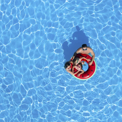 La imagen muestra una vista aérea de una piscina de agua cristalina en la que varias personas se encuentran sobre un flotador circular con diseño de sandía. La composición destaca por el amplio espacio ocupado por el agua azul, donde los reflejos de la luz generan patrones ondulados en toda la superficie. El flotador, de colores rojo, verde y negro, contrasta con el fondo acuático y se sitúa en la zona central de la imagen, convirtiéndose en el punto focal de la escena. En conjunto, la fotografía transmite una experiencia asociada al ocio, el disfrute de instalaciones recreativas y el tiempo libre en un entorno acuático, evocando actividades de descanso y convivencia durante la temporada estival.