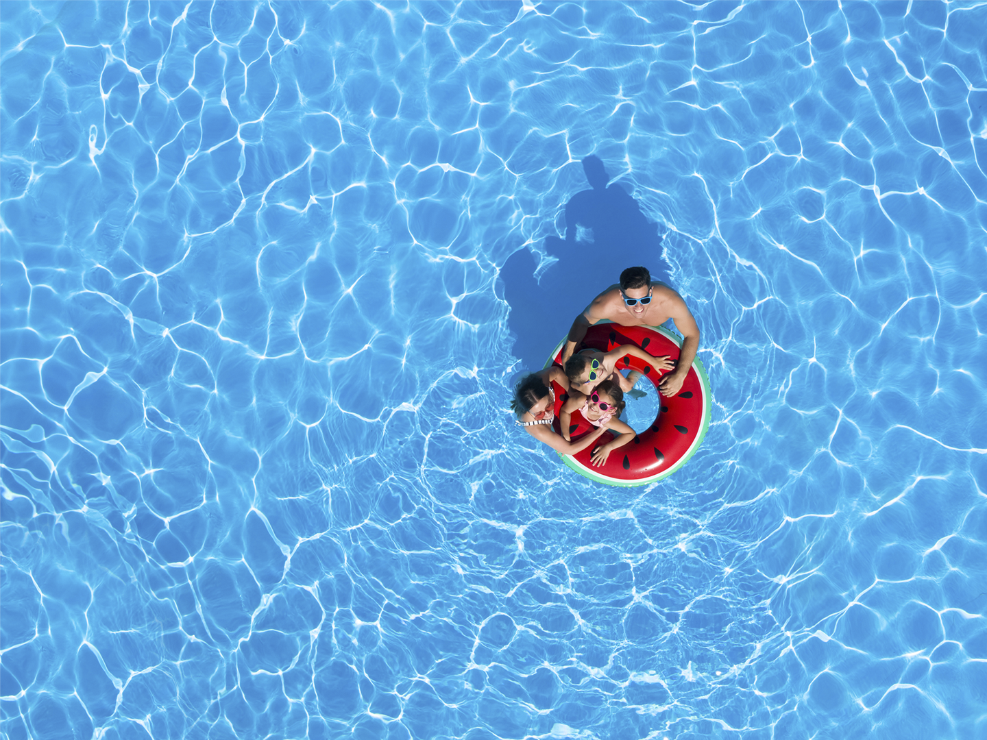La imagen muestra una vista aérea de una piscina de agua cristalina en la que varias personas se encuentran sobre un flotador circular con diseño de sandía. La composición destaca por el amplio espacio ocupado por el agua azul, donde los reflejos de la luz generan patrones ondulados en toda la superficie. El flotador, de colores rojo, verde y negro, contrasta con el fondo acuático y se sitúa en la zona central de la imagen, convirtiéndose en el punto focal de la escena. En conjunto, la fotografía transmite una experiencia asociada al ocio, el disfrute de instalaciones recreativas y el tiempo libre en un entorno acuático, evocando actividades de descanso y convivencia durante la temporada estival.