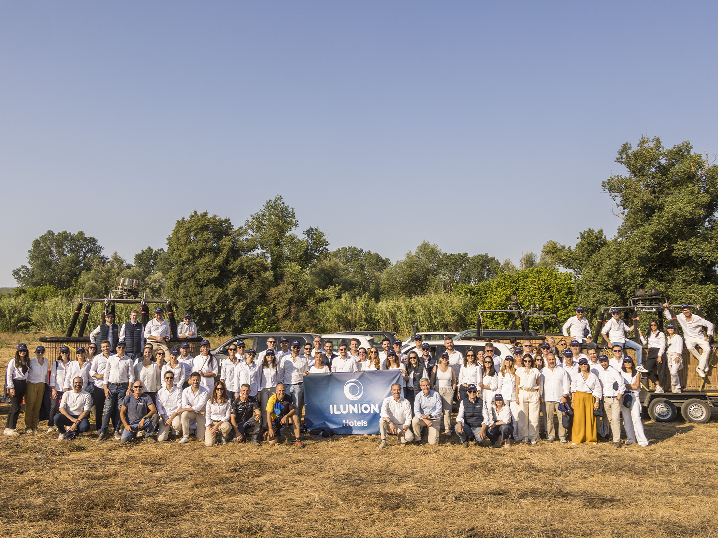Grupo numeroso de personas posando en un campo seco al aire libre, organizadas en varias filas y vestidas mayoritariamente con ropa clara, con una pancarta azul de “ILUNION” en el centro; a ambos lados se observan vehículos tipo remolque con más personas subidas, y al fondo aparecen árboles verdes bajo un cielo despejado de gran amplitud.