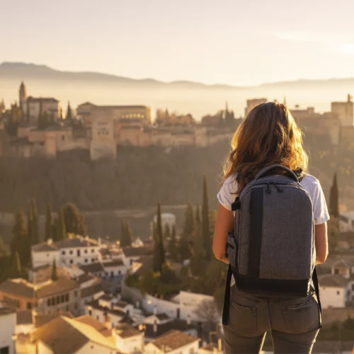 Persona en primer plano vista de espaldas, de pie en un mirador elevado, con una mochila gris de gran tamaño colocada sobre la espalda y vestimenta informal de tonos claros. Frente a la persona se extiende una ciudad histórica con edificaciones compactas, torres y cúpulas, parcialmente envuelta por una luz cálida de atardecer que crea un ambiente dorado y difuso. En el fondo se distinguen colinas suaves y una ligera bruma que atenúa los contornos del paisaje. En la parte inferior aparecen tejados y árboles altos y estrechos que aportan profundidad, mientras la iluminación suave resalta los volúmenes y genera una atmósfera tranquila y contemplativa.