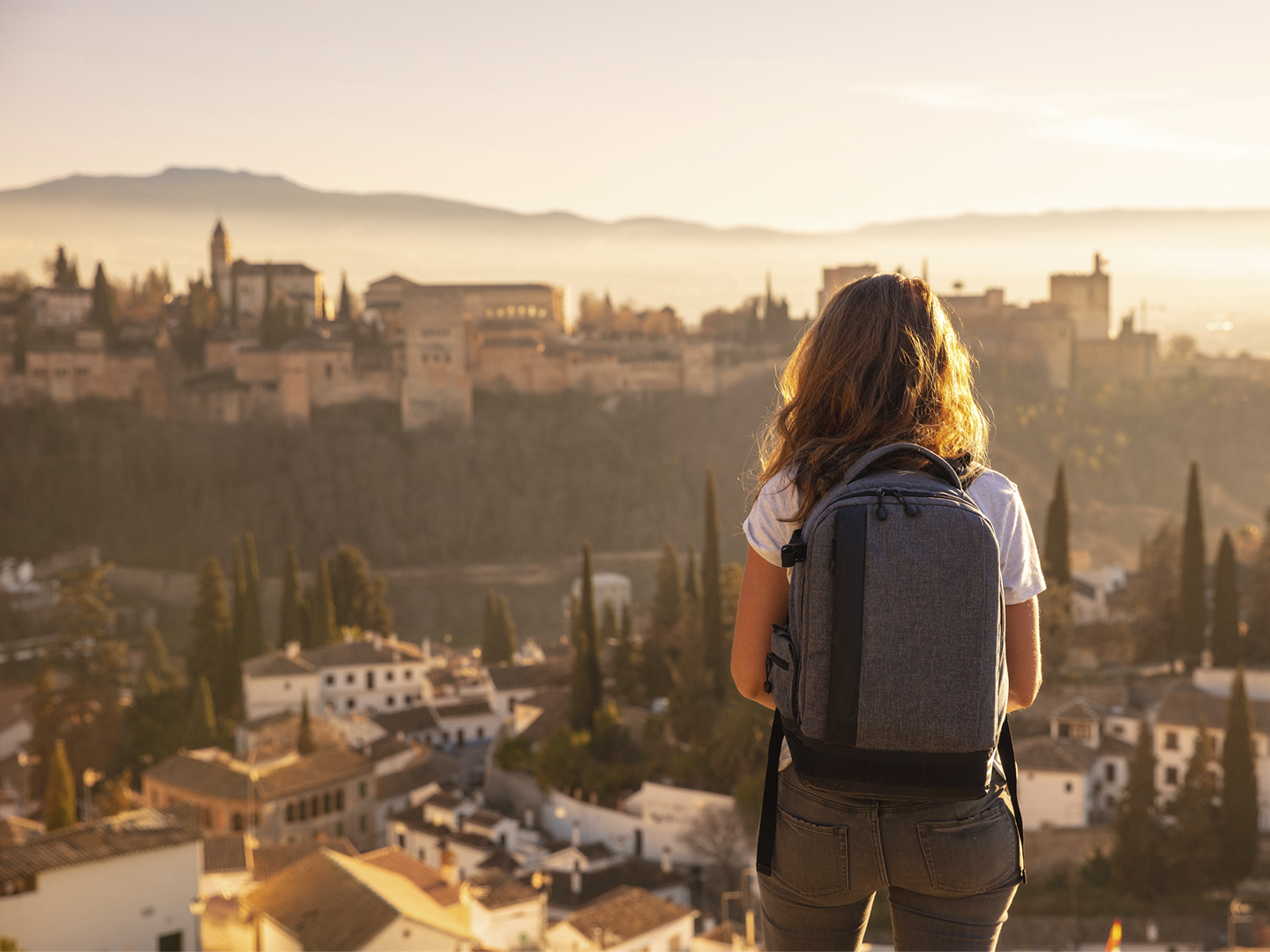 Persona en primer plano vista de espaldas, de pie en un mirador elevado, con una mochila gris de gran tamaño colocada sobre la espalda y vestimenta informal de tonos claros. Frente a la persona se extiende una ciudad histórica con edificaciones compactas, torres y cúpulas, parcialmente envuelta por una luz cálida de atardecer que crea un ambiente dorado y difuso. En el fondo se distinguen colinas suaves y una ligera bruma que atenúa los contornos del paisaje. En la parte inferior aparecen tejados y árboles altos y estrechos que aportan profundidad, mientras la iluminación suave resalta los volúmenes y genera una atmósfera tranquila y contemplativa.