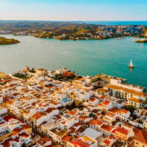 Vista aérea de una localidad costera con casas blancas y tejados rojizos junto a una amplia bahía de aguas turquesa. En el centro del mar navega un velero de vela blanca, mientras al fondo se extienden colinas cubiertas de vegetación y pequeñas urbanizaciones junto al litoral. La escena está iluminada por una luz cálida que resalta los colores del paisaje mediterráneo.