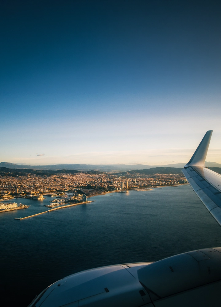 Vista aérea desde un avión de una ciudad costera bañada por luz cálida, con el ala y parte del motor visibles en el lado derecho en primer plano; debajo se extiende un puerto con diques y embarcaciones, seguido de numerosos edificios que se agrupan hasta las montañas del fondo. El mar ocupa gran parte de la escena con un tono azul profundo que contrasta con el cielo despejado en degradado, creando una sensación de amplitud y altura.