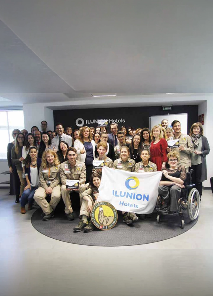 Grupo numeroso posando en el interior de un hotel frente al logotipo de ILUNION Hotels. Varias personas sostienen una bandera de la marca y diplomas, mientras una persona aparece en silla de ruedas en primera fila.