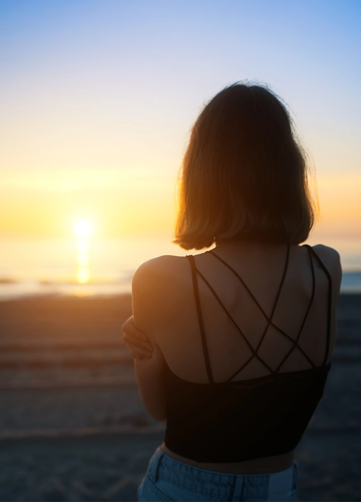 Persona de espaldas contemplando la puesta de sol en la playa. Viste una camiseta de tirantes con la espalda cruzada y se encuentra frente al mar, iluminada por la luz cálida del atardecer. El sol se refleja sobre el agua mientras el cielo muestra una transición suave de tonos dorados a azul.