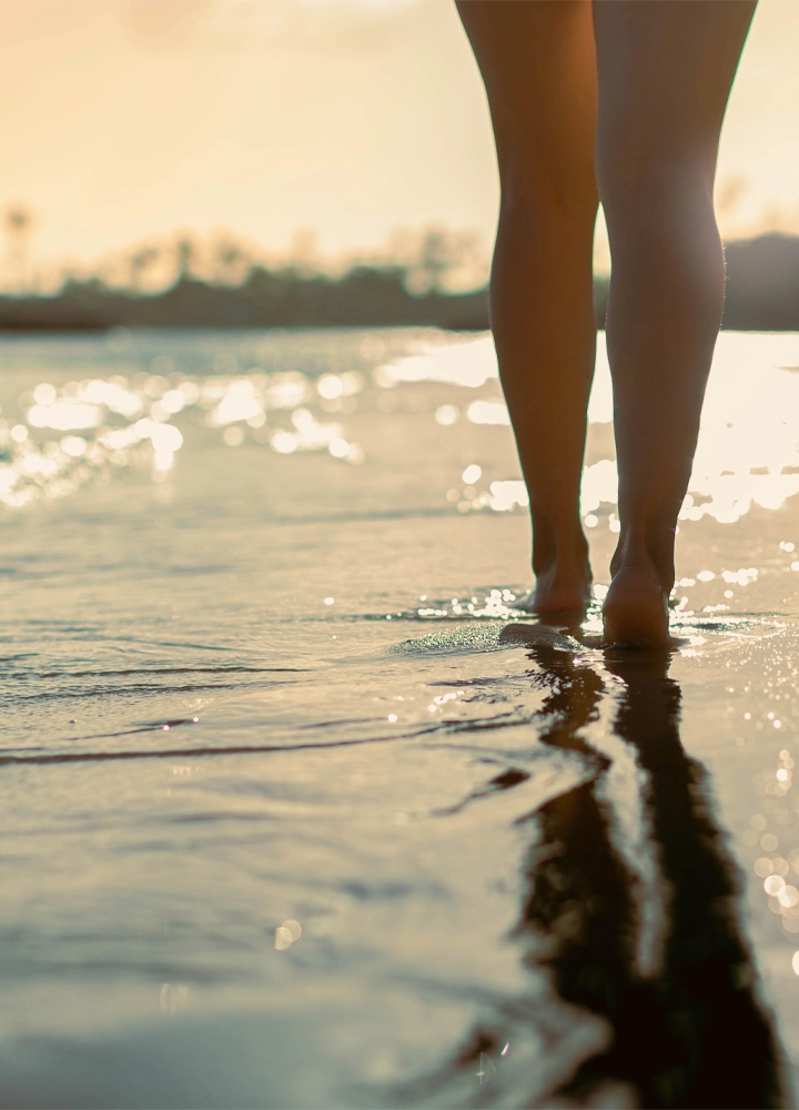 Piernas descalzas caminando por la orilla de una playa con poca profundidad. La luz dorada del atardecer se refleja en el agua y crea destellos sobre la superficie, mientras el reflejo de la figura se alarga en la arena húmeda. El fondo aparece desenfocado, con el horizonte y la vegetación apenas visibles.