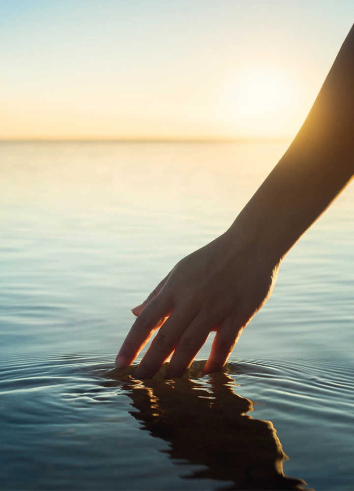 Mano rozando suavemente la superficie de un agua tranquila al atardecer. Los dedos crean pequeñas ondas concéntricas mientras la luz dorada del sol se refleja sobre el agua, proyectando una silueta y un reflejo alargado. El horizonte aparece difuminado bajo un cielo despejado en tonos cálidos y suaves.
