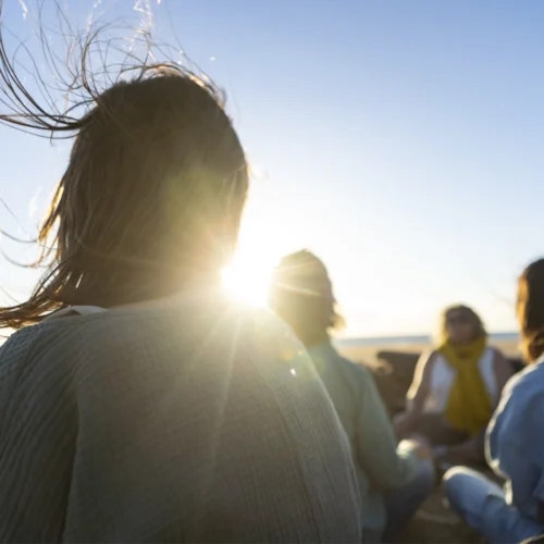 Grupo de personas reunidas en una playa al atardecer. En primer plano aparece una persona de espaldas, con el cabello movido por el viento y el sol brillando entre los mechones. Al fondo, varias personas están sentadas en círculo cerca de la orilla, iluminadas por la luz cálida del sol bajo sobre el horizonte.